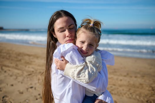 Caucasian delightful young mother gently hugging her little kid girl, standing with her eyes closed on the sandy beach, against the background of beautiful waves pounding on the Atlantic Sea shore
