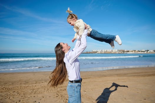 Happy mother holding her little child, adorable daughter girl in her outstretched hands, throwing up and playing on the nature, standing together on the sandy beach against crashing waves background