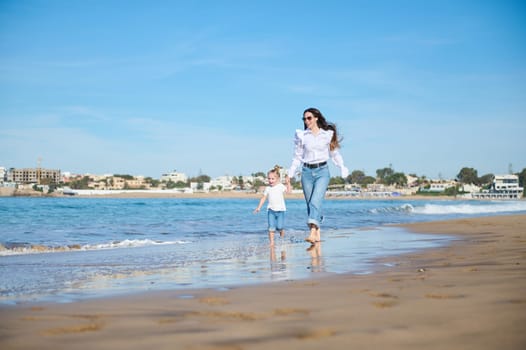 Mother and daughter holding hands, walking together barefoot on the splashing waves of tropical sandy beach on warn sunny day, enjoying happy moments together. People. Lifestyle. Leisure activity