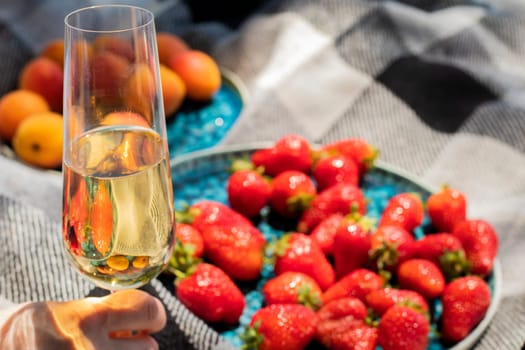 strawberries on plate with glass of champagne or white wine on picnic. Luxury lifestyle, travel concept. glass raised in hand for toast. Ripe berries. wine tasting. Vineyard. Summer fruits. aesthetics