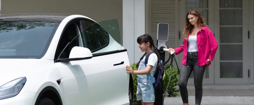 Happy little young girl learn about eco-friendly and energy sustainability as she help her mother recharge electric vehicle from home EV charging station. EV car and modern family. Panorama Synchronos