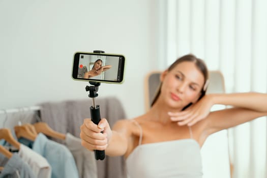 Young woman making fashion video content for social media. Blogger smiles to camera using selfie stick and light ring while making persuasive online clothing sell to audience or followers. Unveiling