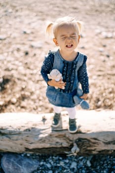 Little girl grimaces while standing on a snag with toys on the beach. High quality photo