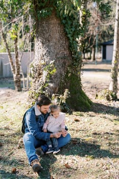 Little girl sits on her father lap on the grass under a large tree, looking at a fir cone in his hands. High quality photo