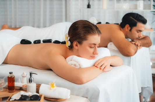 Hot stone massage at spa salon in luxury resort with day light serenity ambient, blissful couple customer enjoying spa basalt stone massage glide over body with soothing warmth. Quiescent