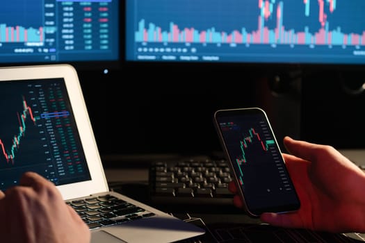 Cropped image of young trader searching on smartphone comparing with laptop and dynamic financial exchange rate data screens of current market graph online in real time at neon modern office. Gusher.