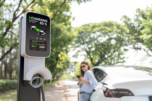 Young woman using smartphone online banking application to pay for electric car battery charging from EV charging station during vacation holiday road trip at national park or summer forest. Exalt