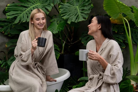 Tropical and exotic spa garden with bathtub in modern hotel or resort with young two women in bathrobe drink coffee, enjoy leisure and wellness lifestyle surround by lush greenery foliage. Blithe