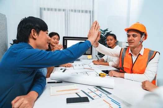 Diverse group of civil engineer and client celebrate and high five after make successful agreement on architectural project, reviewing construction plan and building blueprint at meeting table.Prudent