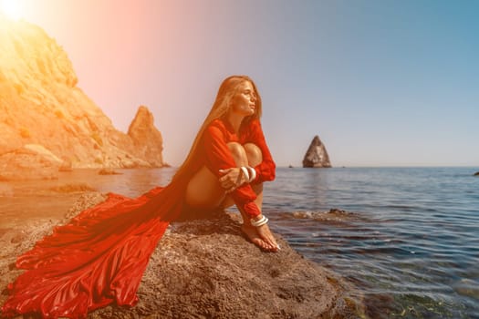 woman sea red dress. Beautiful sensual woman in a flying red dress and long hair, sitting on a rock above the beautiful sea in a large bay