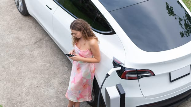 Aerial top view eco-friendly woman recharge electric vehicle from EV charging station, using EV technology utilization for tracking energy usage to optimize battery charging on smartphone. Synchronos