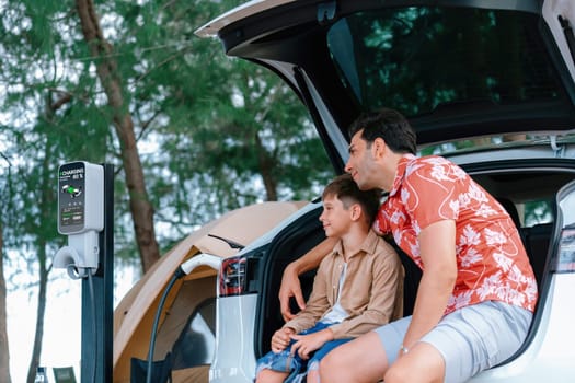 Outdoor adventure and family vacation camping in nature travel by eco friendly car for sustainable future. Father and son sit on trunk, charging EV car with EV charging station in campsite. Perpetual