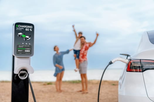 Alternative family vacation trip traveling by the beach with electric car recharging battery from EV charging station with blurred cheerful and happy family enjoying the seascape background. Perpetual