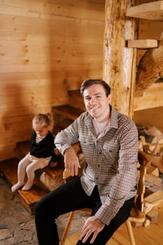 Smiling dad sitting on chair next to little girl sitting on stairs in wooden house. High quality photo