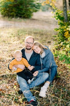 Mom rested her head on the shoulder of dad sitting with his little son on his knees on dry grass. High quality photo