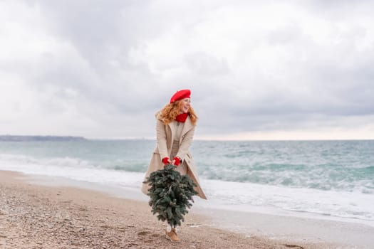 Redhead woman Christmas tree sea. Christmas portrait of a happy redhead woman walking along the beach and holding a Christmas tree in her hands. She is dressed in a light coat and a red beret