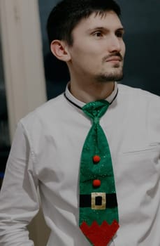 Portrait of one young handsome brunette guy in a white shirt with a green Christmas Santa Claus tie looking to the side, close-up side view.
