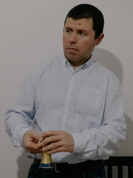 Portrait of one young Caucasian man in a blue shirt, standing at a festive table and thoughtfully opening a bottle of champagne, looking to the side, close-up side view.