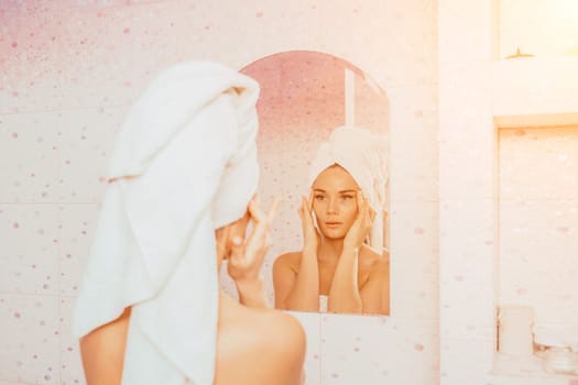 Young beautiful woman using face cream moisturizing lotion after bath. Pretty attractive girl in a towel on her head stands in front of a mirror in a home bathroom. Daily hygiene and skin care.