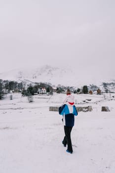 Father with a small child on his shoulders walks along a snow-covered hill towards a village at the foot of the mountains. Back view. High quality photo