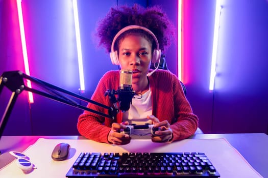 Host channel of gaming streamer, African girl playing online game with joystick, talking with viewers media online on microphone. Esport skilled team players in neon color lighting room. Tastemaker.