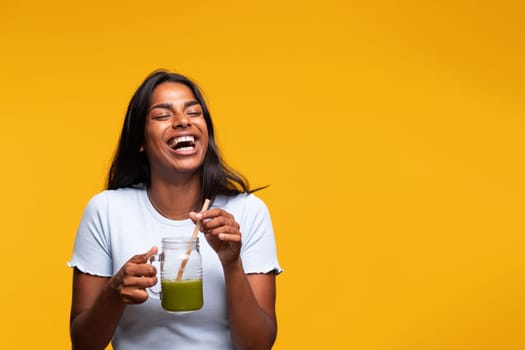 Happy young beautiful Indian woman with green juice on yellow background, laughing. Copy space. Healthy lifestyle concept.