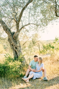 Guy and girl are sitting hugging on the grass under a tree with their noses touching. High quality photo