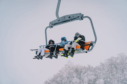 Skiers on a chairlift ride uphill above snow-covered trees. High quality photo