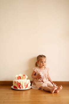 Little girl with a doll in her hand sits on the floor next to a birthday cake on a plate. High quality photo