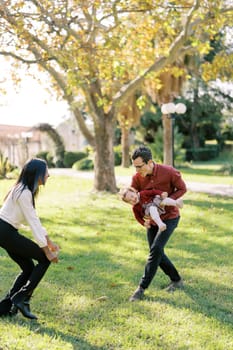 Dad with a little girl in his arms goes crouching to a laughing mom in the park. High quality photo