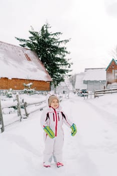 Little smiling girl stands on a snowy road near a fence in the village. High quality photo