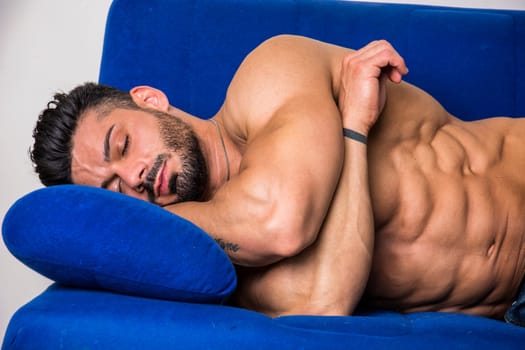 Muscular shirtless bodybuilder sleeping on blue sofa wearing only jeans