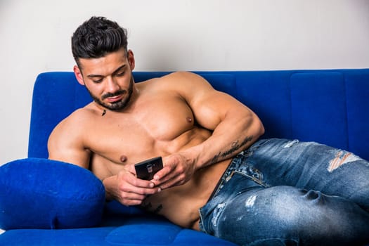 Handsome shirtless muscular bodybuilder man typing or surfing the internet with cell phone while laying on couch