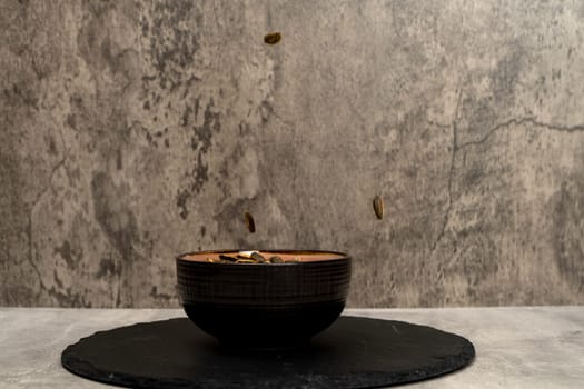 Sunflower seeds falling into a ceramic bowl with more salted sunflower seeds on a gray background.