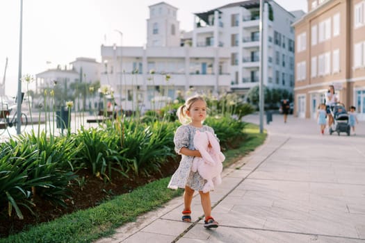 Little girl with a pink plush rabbit in her hands stands on a path in the courtyard of a multi-story building. High quality photo