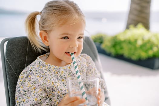 Little girl bites a striped straw in a glass of water in her hands while sitting in a chair. High quality photo