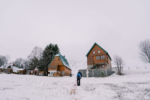 Small child drags a sled on a rope to his dad standing on a hill near a wooden cottage. Back view. High quality photo