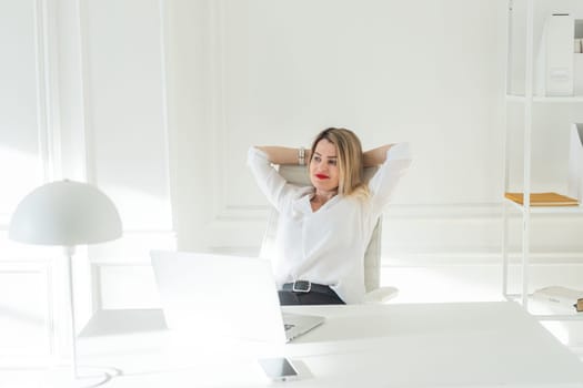 Delighted young blonde woman sitting at white table in blouse using laptop. High quality photo