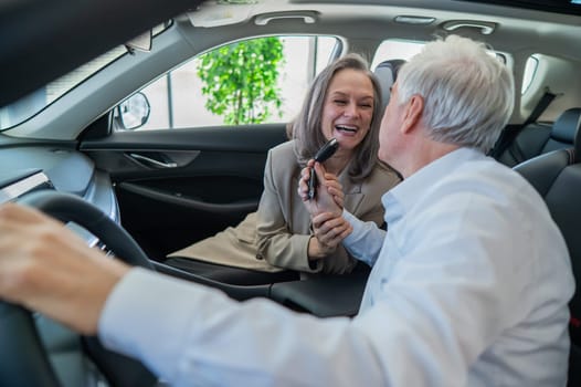 An elderly man holds the keys while sitting in a new car. A gray-haired married couple is happy about buying a car