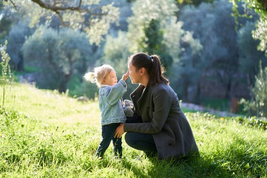 Little girl touches her mom nose while squatting on a green lawn. High quality photo