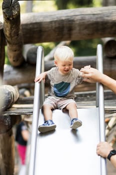 Parent hand holding little infant baby boy child while sliding on urban playground on a sunny summer day. Family joy and happiness concept.