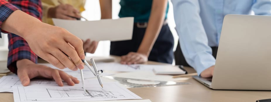 Architect team discuss about blueprint design at modern meeting room while her coworker working on laptop on table with blueprint and architectural equipment scattered around. Closeup. Delineation.