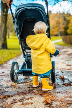Sun always shines after the rain. Small blond infant boy wearing yellow rubber boots and yellow waterproof raincoat walking in puddles, pushing stroller in city park, holding mother's hand after rain