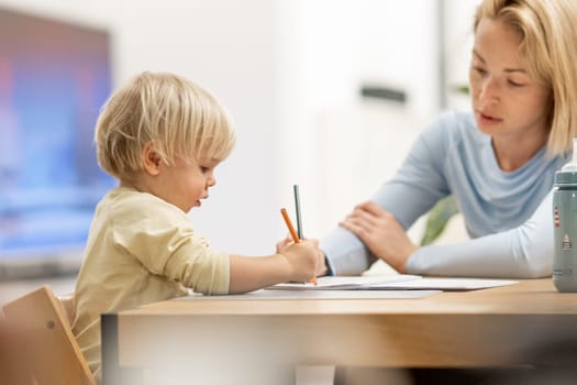 Caring young Caucasian mother and small son drawing painting in notebook at home together. Loving mom or nanny having fun learning and playing with her little 1,5 year old infant baby boy child