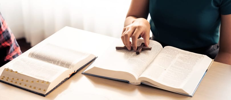 Close-up female Christian prayer pickup holy cross on bible book at wooden church table while asian believer reading bible. Concept of religion, faith, and god blessing. Warm background. Burgeoning.