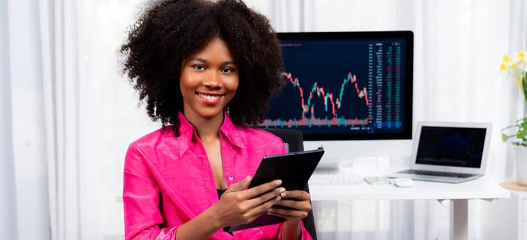 Analytical young African American businesswoman, a specialist in successful stock exchange trading, against dynamic data graph displaying marketing trend analysis on screen. Tastemaker.