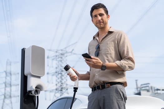 Man pay for electricity with smartphone while recharge EV car battery at charging station connected to power grid tower electrical as electrical industry for eco friendly car utilization.Expedient