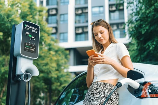 Young woman travel with EV electric car charging in green sustainable city outdoor garden in summer. Urban sustainability lifestyle by green clean rechargeable energy of electric BEV vehicle innards