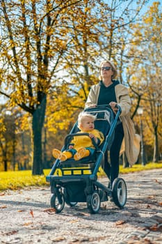 Young beautiful mother wearing a rain coat pushing stroller with her little baby boy child, walking in city park on a sunny autumn day.