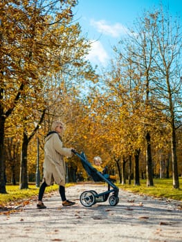 Young beautiful mother wearing a rain coat pushing stroller with her little baby boy child, walking in city park on a sunny autumn day.